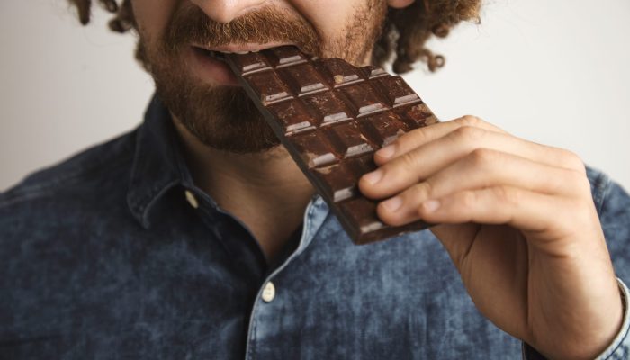 Closeup curly hair happy bearded man with healthy skin bites organic freshly baked chocolate bar with side of mouth, close focus on mouth